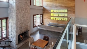 Un interior espacioso con chimenea de piedra, pared de madera y barandilla de vidrio con vista a un nivel inferior.