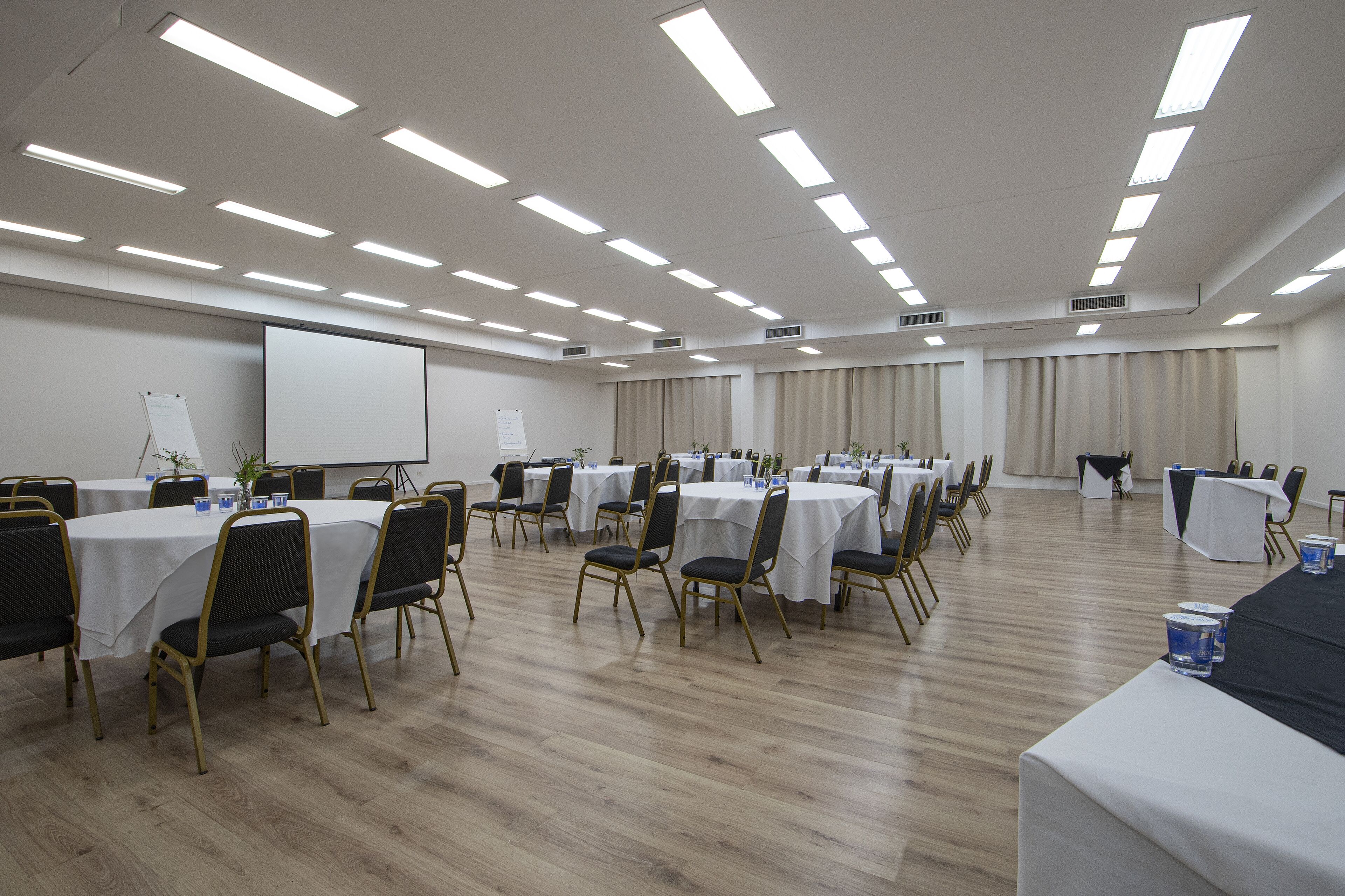 Sala de conferências com várias mesas e cadeiras, paredes brancas e luminárias no teto.