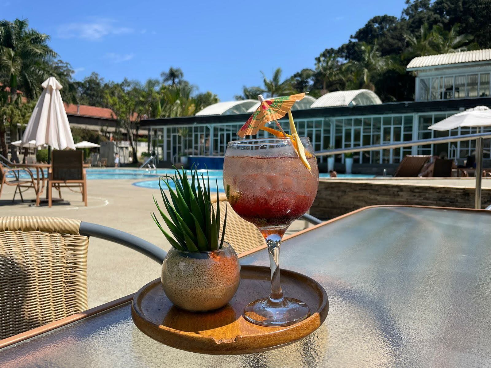 Cóctel sobre una bandeja junto a una piscina con sillones de descanso y sombrillas al fondo.