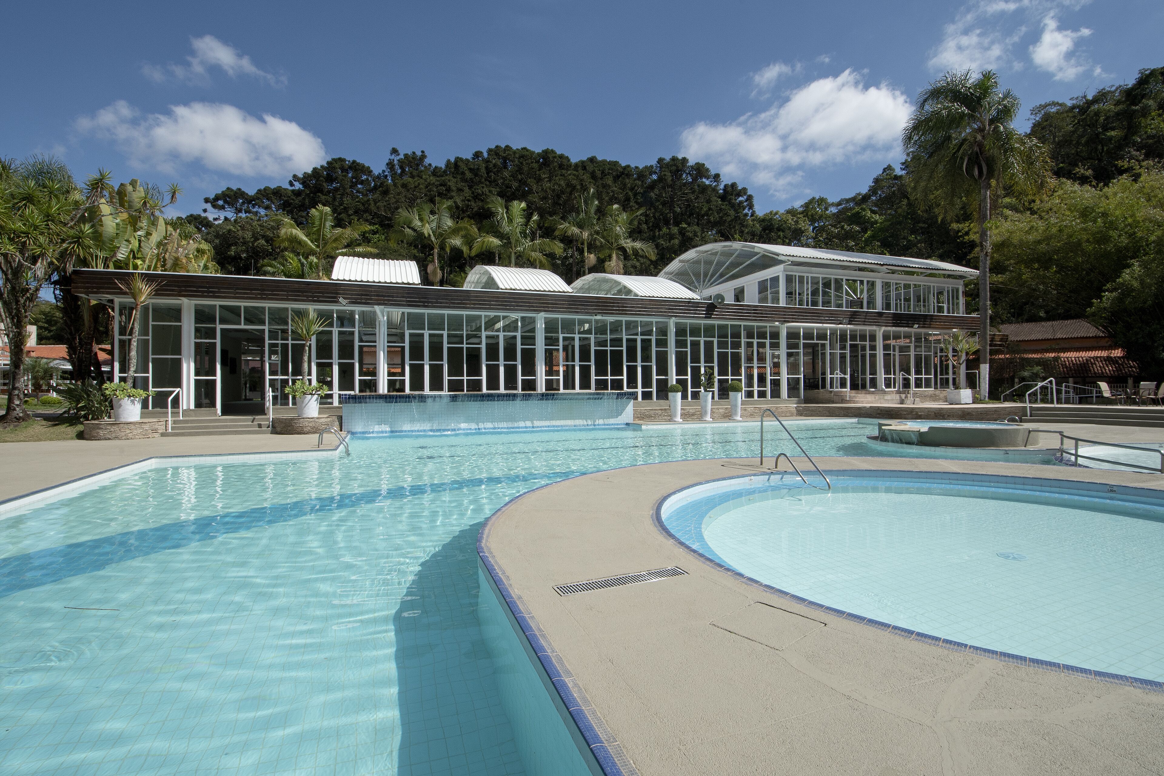 Ein großer Außenpool mit klarem, blauem Wasser, umgeben von einer Glas-Orangerie, Palmen und einem wolkenlosen Himmel.