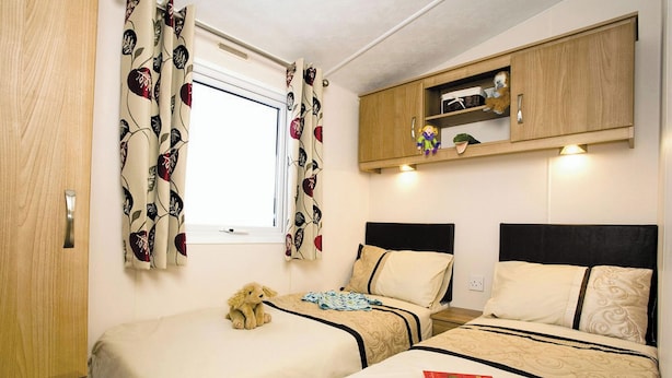 A room with two beds, a window with floral curtains, a wooden cabinet above the beds, and a teddy bear on one bed.