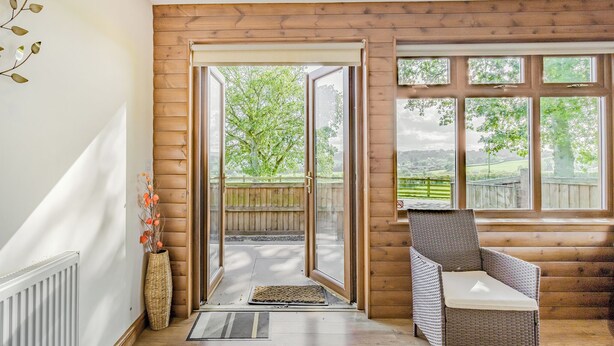 A room with a wooden floor, a wicker chair, a door leading outside, and a window with a view of greenery. - Valley View Lodge Pet (Penybont, Llandrindod Wells)