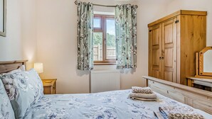 A bedroom with a bed, a nightstand, a wooden wardrobe, and a window with floral curtains. - Valley View Lodge Pet (Penybont, Llandrindod Wells)