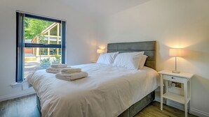 Een netjes opgemaakt bed met witte lakens en handdoeken, een nachtkastje met een lamp, en een raam met uitzicht op groen.