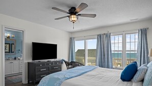 A bedroom with a bed, a TV, a ceiling fan, a dresser, and a window with curtains. - Oceanfront duplex with direct beach access and community pool, Spinnakers Reach (Emerald Isle)