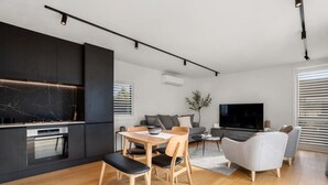 A modern kitchen with dark cabinetry and a marble backsplash, a dining area with wooden furniture, a grey sofa, and a flat-screen TV in a living room with large windows. - Nestled below the Remarkables (Jacks Point)