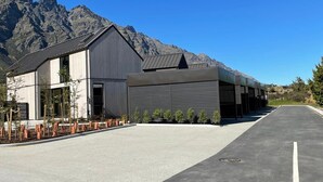 A modern building with a flat roof and large windows, surrounded by a paved area with a clear sky and mountains in the background. - Nestled below the Remarkables (Jacks Point)