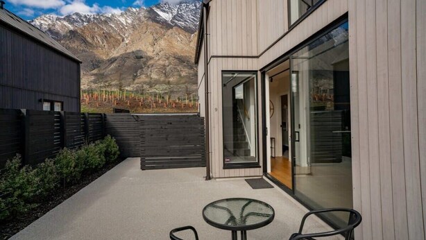 A modern building with a glass door, a patio with a table and chairs, and a mountainous backdrop. - Nestled below the Remarkables (Jacks Point)