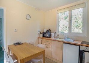 A compact kitchen with a wooden table, white chairs, a microwave, kettle, and a sink. - Comfortable log cabin offering self-catering in the heart of Cornwall. (Ruthernbridge, Nr Bodmin)