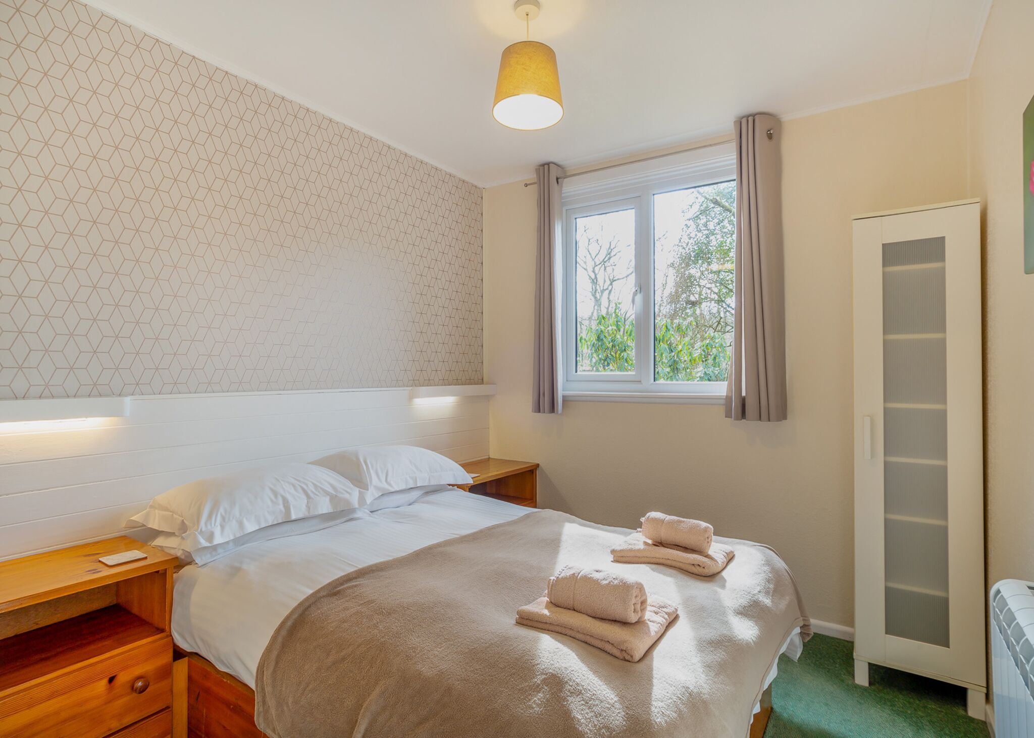Une chambre d’hôtel comprenant un lit, des tables de chevet, une fenêtre avec des rideaux, une armoire et un mur orné d’un motif géométrique.