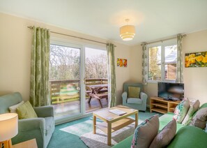 A living room with a sofa, armchair, coffee table, TV, and a sliding glass door leading to an outdoor deck. - Comfortable log cabin offering self-catering in the heart of Cornwall. (Ruthernbridge, Nr Bodmin)