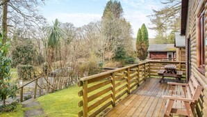 A wooden deck with a picnic table and benches, surrounded by trees and a view of a cabin. - Comfortable log cabin offering self-catering in the heart of Cornwall. (Ruthernbridge, Nr Bodmin)