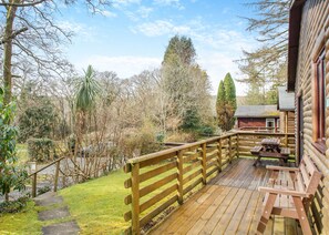 A wooden deck with a picnic table and benches, surrounded by trees and a view of a cabin. - Comfortable log cabin offering self-catering in the heart of Cornwall. (Ruthernbridge, Nr Bodmin)