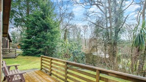 A wooden deck with a railing, a bench, and a view of a forested area with trees and a small cabin. - Comfortable log cabin offering self-catering in the heart of Cornwall. (Ruthernbridge, Nr Bodmin)