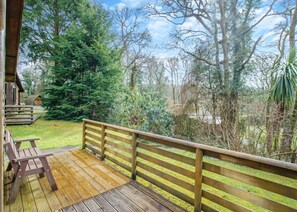 A wooden deck with a railing, a bench, and a view of a forested area with trees and a small cabin.
