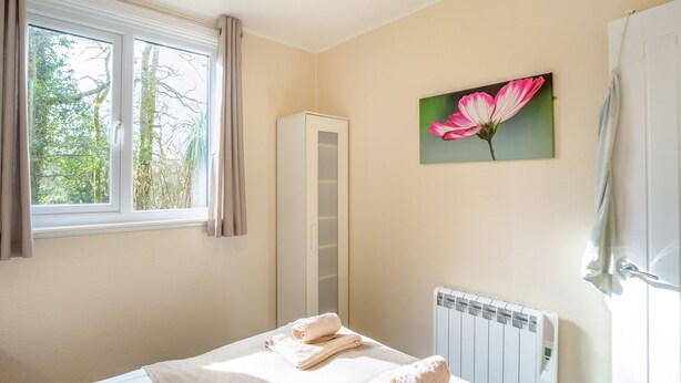 A bedroom with a bed, a radiator, a window with curtains, and a painting of a flower. - Comfortable log cabin offering self-catering in the heart of Cornwall. (Ruthernbridge, Nr Bodmin)