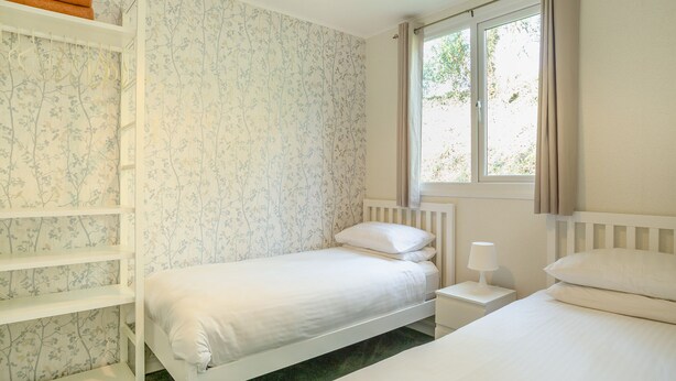 A bedroom with two beds, a window with curtains, a radiator, and a shelf with towels. - Comfortable log cabin offering self-catering in the heart of Cornwall. (Ruthernbridge, Nr Bodmin)