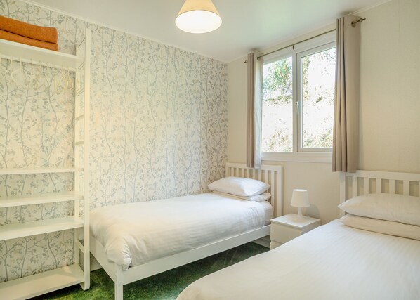 A bedroom with two beds, a window with curtains, a radiator, and a shelf with towels. - Comfortable log cabin offering self-catering in the heart of Cornwall. (Ruthernbridge, Nr Bodmin)