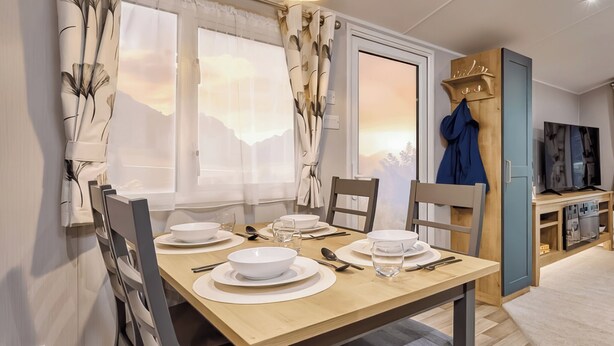 Un área de comedor con una mesa de madera preparada para cuatro personas, un televisor sobre un mueble de madera y vistas a montañas a través de las ventanas.