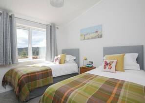 A bedroom with two beds, a window with curtains, a bedside table with a lamp, and a painting on the wall.