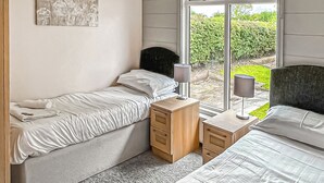 A bedroom with two beds, a nightstand, a lamp, and a window with a view of greenery. - Luxury lodge with one double with en suite shower and one twin. (Thirsk)
