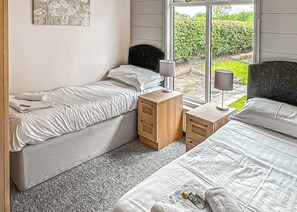 A bedroom with two beds, a nightstand, a lamp, and a window with a view of greenery.