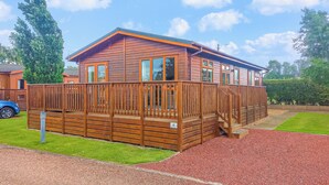 A wooden cabin with a deck, a gravel driveway, and a green lawn. - Luxury lodge with one double with en suite shower and one twin. (Thirsk)