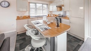 A compact kitchen with a central island, a sink, and a stove. There are two bar stools and a clock on the wall. - Luxury lodge with one double with en suite shower and one twin. (Thirsk)