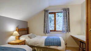 Ein Schlafzimmer mit Bett, Nachttisch, Lampe, Fenster mit Vorhängen, Heizkörper und einer kleinen Schreibtischzeile.