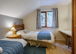 Ein Schlafzimmer mit Bett, Nachttisch, Lampe, Fenster mit Vorhängen, Heizkörper und einer kleinen Schreibtischzeile.