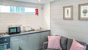 Et tekøkken med mikroovn, brødrister og gaskogeplade. Der er en grå sofa med lyserøde puder og et lille bord med stole.