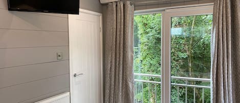 A room with a television mounted on the wall, a white door, a window with curtains, and a radiator.