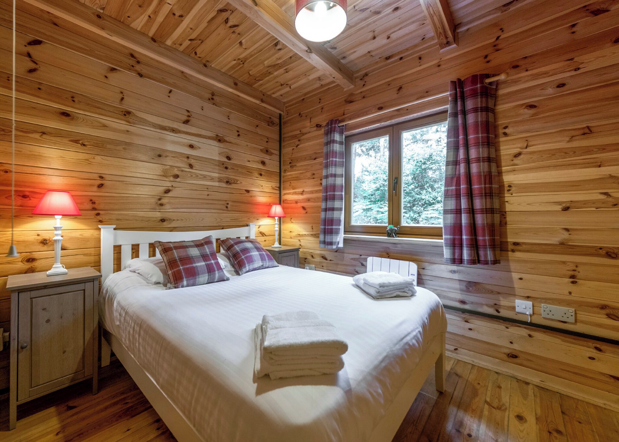 A wooden cabin bedroom with a bed, two bedside lamps, a window with curtains, and a small shelf by the window.