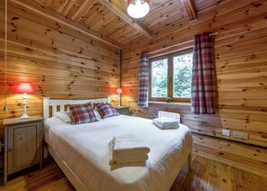 A wooden cabin bedroom with a bed, two bedside lamps, a window with curtains, and a small shelf by the window.