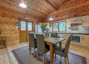 A wooden interior with a dining table, chairs, a refrigerator, a washing machine, and a window with a view of trees.