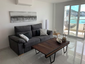 A modern living room with a grey sofa, a wooden coffee table, and a large window offering a view of the sea.