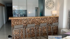 A modern kitchen with a wooden bar area, a large wall clock, and a minimalist design. - Santa Ponsa Mallorca first line of sea - Aparthotel (Santa Ponça)
