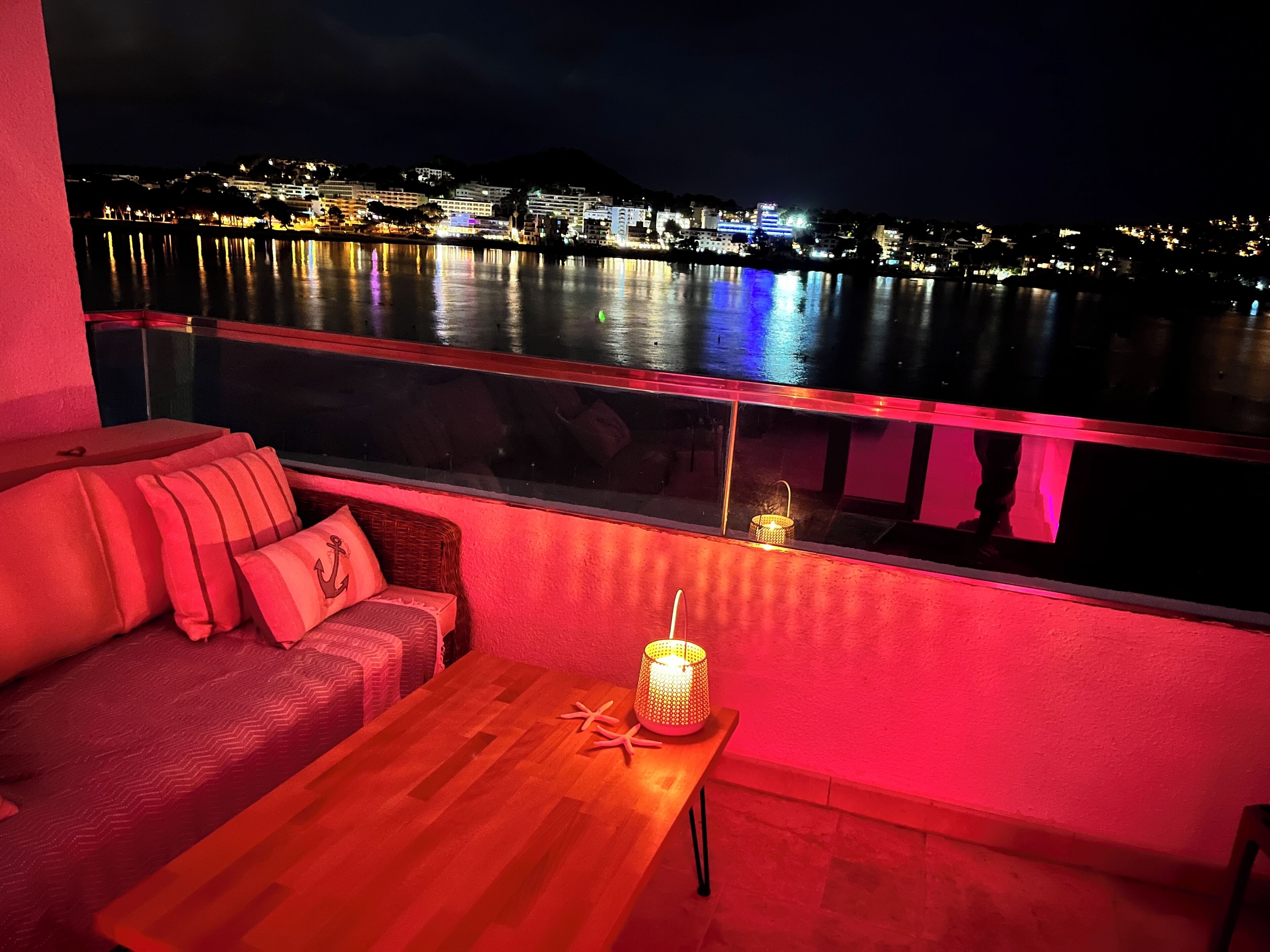 Un balcón con mesa de madera, cojines rojos y sofá de mimbre con vista al frente marítimo de noche.