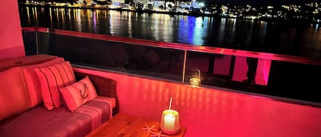 Un balcón con mesa de madera, cojines rojos y sofá de mimbre con vista al frente marítimo de noche.