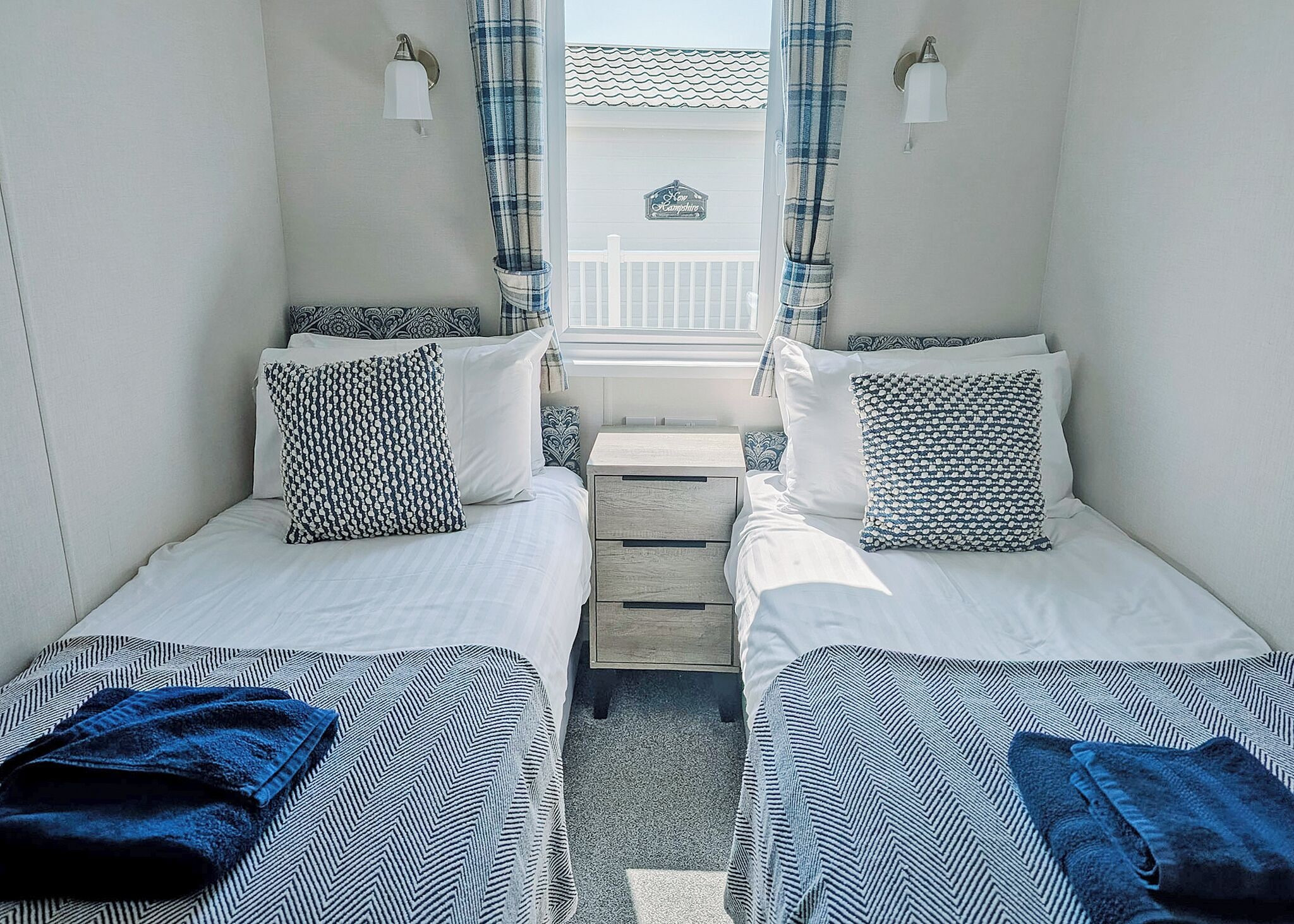 A room with two single beds, each with patterned pillows and a blue towel. There are two wall-mounted light fixtures and a small wooden nightstand between the beds.