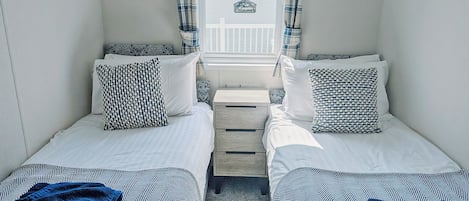 A room with two single beds, each with patterned pillows and a blue towel. There are two wall-mounted light fixtures and a small wooden nightstand between the beds.