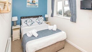 Een slaapkamer met een bed, twee nachtkastjes met lampen, een raam met gordijnen, een wandtelevisie en een poster van een strand.