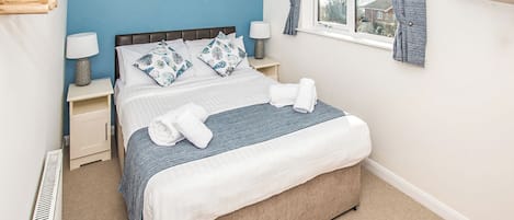 Un dormitorio con una cama, dos mesitas de noche con lámparas, un ventanal con cortinas, un televisor montado en la pared y un cartel con una escena de playa.