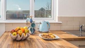 Een keuken met een houten aanrecht, een schaal met fruit, een bord met koekjes, een blauwe waterkoker en een raam met uitzicht op huizen.