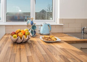 A kitchen with a wooden countertop, a bowl of fruit, a plate of cookies, a blue kettle, and a window with a view of houses.