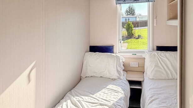 A small, single-bed room with a window, a bedside table, and a power outlet. - York Supeior 3 Caravan (Thirsk)