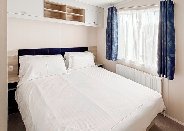 A neatly made bed with white linens, a headboard, and a window with blue curtains.