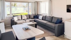 A modern living room with a sectional sofa, a dining table, and a kitchen area. - York Supeior 3 Caravan (Thirsk)