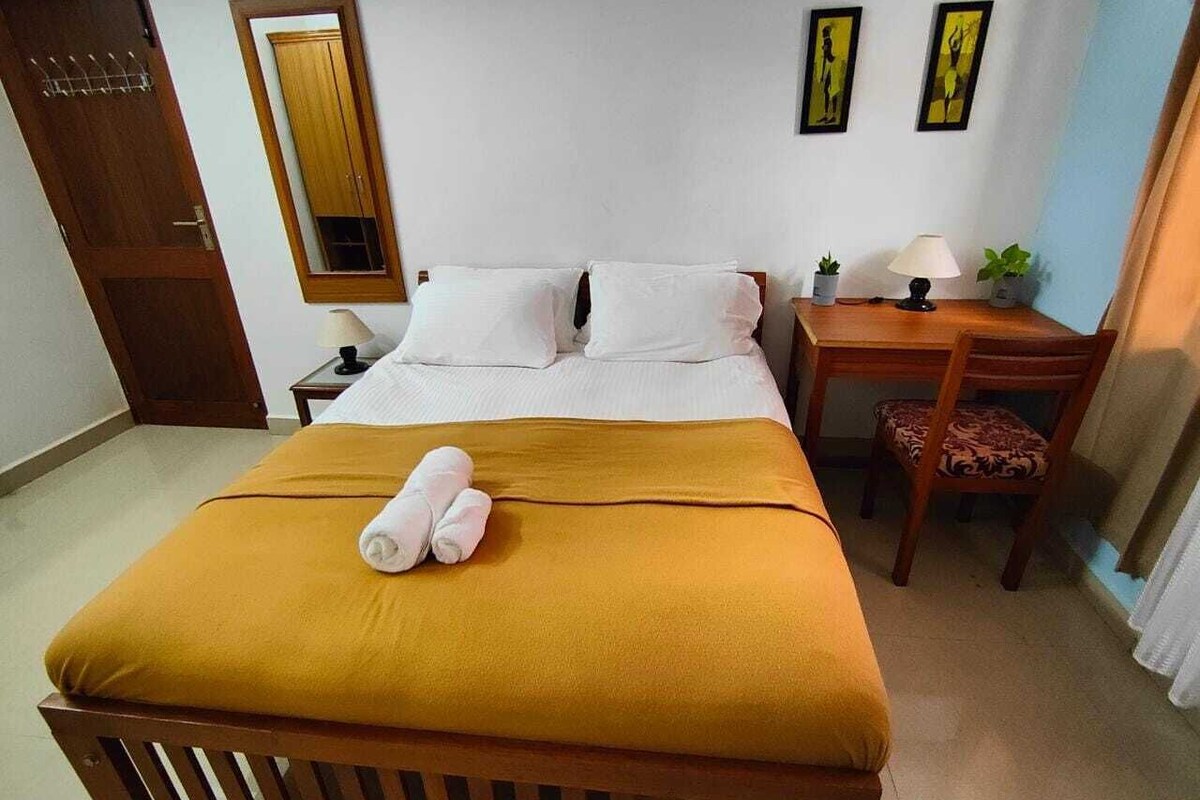 A neatly made bed with a mustard-colored blanket and two rolled towels, a wooden nightstand with a lamp and a small plant, and a wooden door with a mirror.