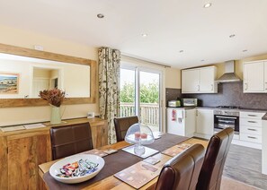 A dining area with a table, chairs, a vase with flowers, a view of the outdoors, and a kitchen with appliances. - Luxury lodge with one double and one twin. (Pickering)